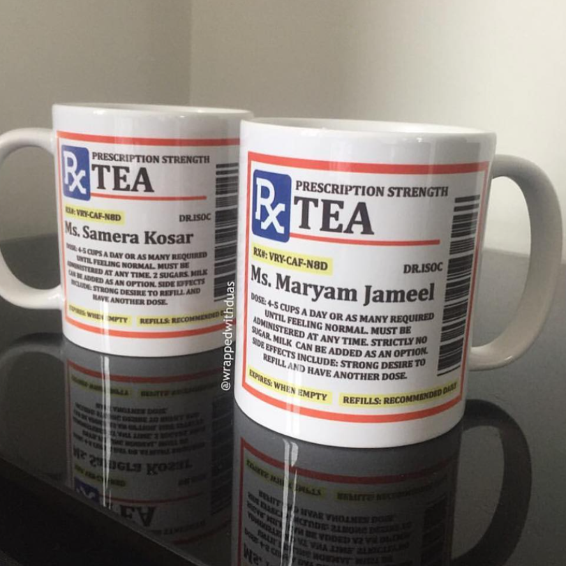 Two white ceramic mugs printed with a parody prescription-strength tea label design, shown on a reflective dark surface against a light wall.