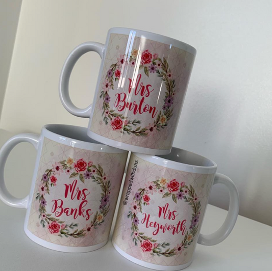 Three ceramic mugs stacked in a pyramid arrangement on a white surface against a light beige wall. Each mug features a floral wreath design with the personalized name of a woman printed in red script: 'Mrs Banks', 'Mrs Heywoods', and 'Mrs Bunten'.