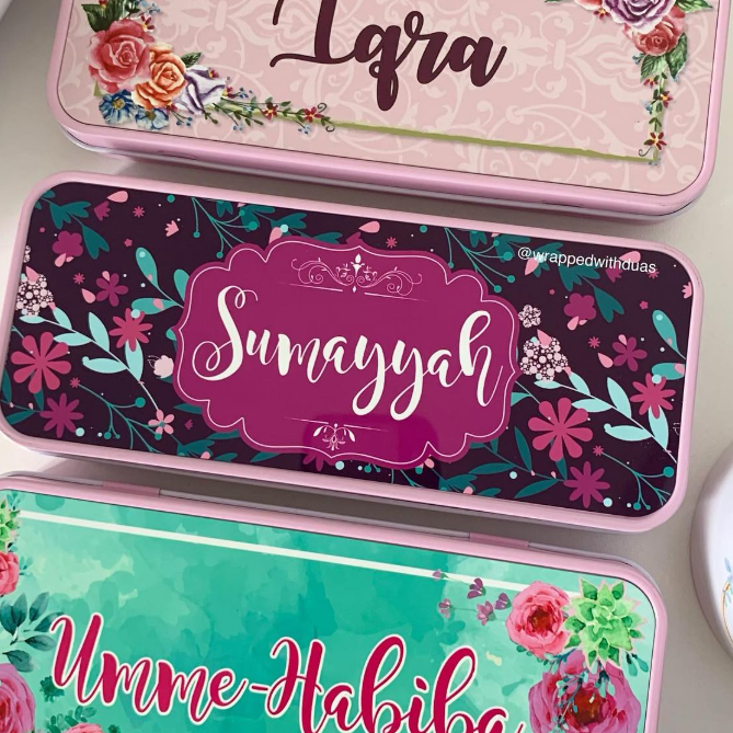 Three personalized metal gift boxes stacked diagonally, each with a different floral design and a custom name: 'Lara' on a pink damask box, 'Sumayah' on a dark purple floral box, and 'Umme-Habiba' on a teal floral box.