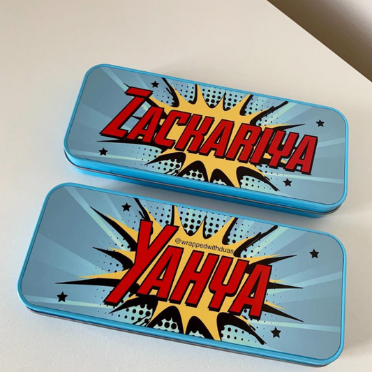 Two blue metal pencil cases with comic-style burst graphics and large red text reading 'Zachariya' and 'Yahya' on a white surface.