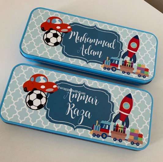 Two personalized metal pencil cases with a blue and white geometric pattern, featuring cartoon illustrations of a red car on a soccer ball, a rocket ship, and a toy train. The top case is personalized with the name 'Muhammad Adam' and the bottom with 'Anwar Raza'.