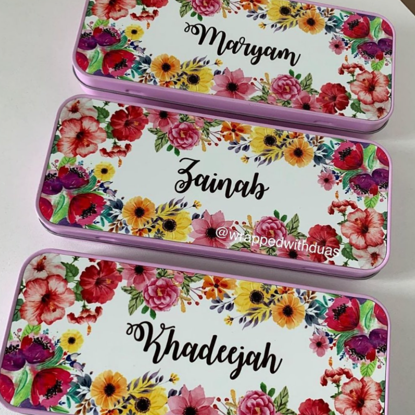 Three rectangular metal gift boxes with floral designs are arranged diagonally on a white surface. The boxes have pink borders, a colorful watercolor floral pattern with red, pink, yellow, and orange flowers, and the names Maryam, Zainab, and Khadeejah written in black script on the top.