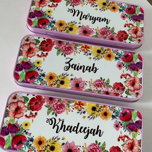 Three rectangular metal gift boxes with floral designs are arranged diagonally on a white surface. The boxes have pink borders, a colorful watercolor floral pattern with red, pink, yellow, and orange flowers, and the names Maryam, Zainab, and Khadeejah written in black script on the top.