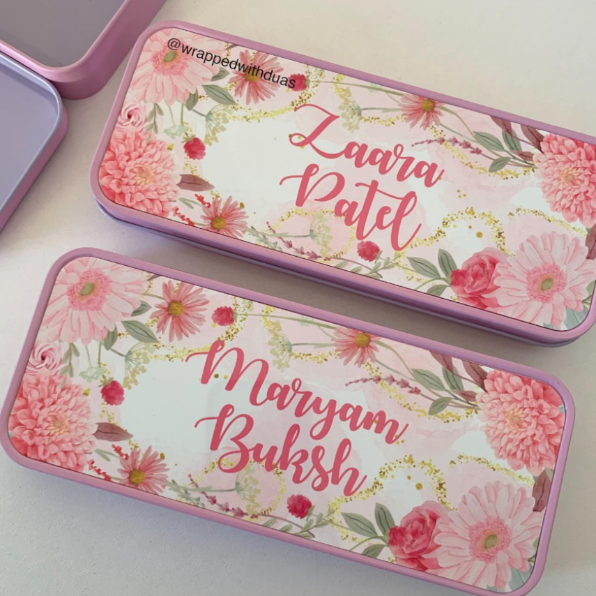Two personalized metal pencil cases with pink floral designs and gold foil accents, shown on a light beige surface. One case is open, revealing a lavender interior, and the other is closed with the name 'Laara Patel' written in pink script.
