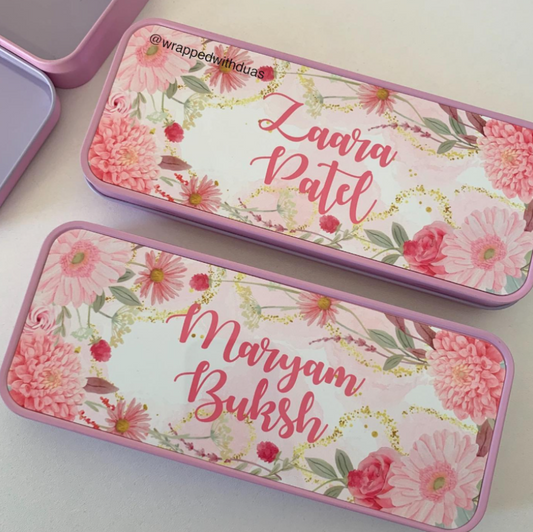 Two personalized metal pencil cases with pink floral designs and gold foil accents, shown on a light beige surface. One case is open, revealing a lavender interior, and the other is closed with the name 'Laara Patel' written in pink script.