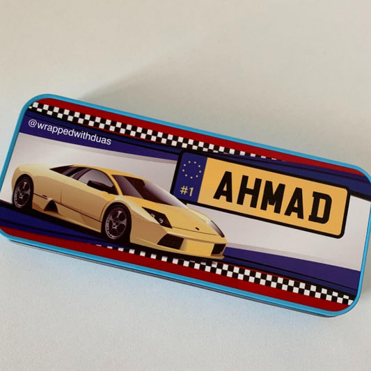 A rectangular metal tin with rounded corners, featuring a printed design of a yellow sports car, a blue and red checkered stripe border, a yellow license plate reading AHMAD, and the handle @wrappedwithduas on the side. The tin is photographed on a plain light background.