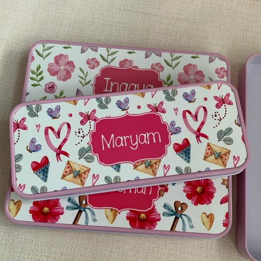 Three personalized rectangular metal gift boxes with pink floral prints and pink borders, stacked on a beige fabric surface. The top box is partially visible, the middle box is labeled 'Maryam', and the bottom box has a floral pattern with hearts, flowers, and ribbon motifs.