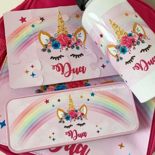 A pink school bag with matching printed accessories, including a puzzle, a bottle, and a pencil case, all featuring a unicorn face, rainbow, flowers, and the name 'Dua'.