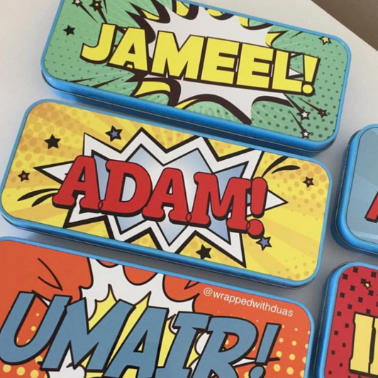 A set of custom comic-style pencil cases arranged on a light surface. The visible cases feature bold pop-art text and graphics: one says 'JAMEEL!' in yellow on green, one says 'ADAM!' in red on yellow, one says 'UMAIR!' in blue on orange