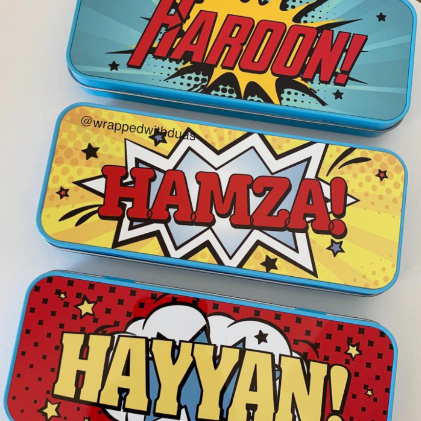 Three comic-style metal pencil cases stacked vertically on a white surface. The top case is blue with red 'HAROOM!' text, the middle is yellow with red 'HAMZA!' text, and the bottom is red with yellow 'HAYYAN!' text.
