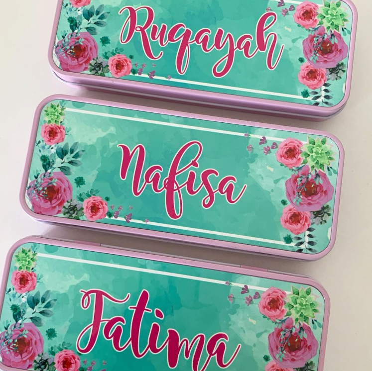 Three rectangular metal nameplates or name tags with rounded corners, arranged vertically on a light background. Each tag has a turquoise floral design with pink roses, green leaves, and a pink border, and the names 'Rugayah', 'Nafisa', and 'Fatima' are written in pink cursive script.