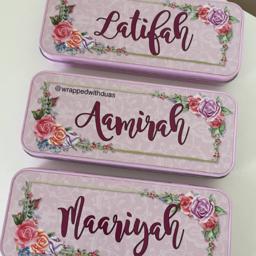 Three personalized pink metal nameplates or nameplates-style cases arranged vertically on a white surface. Each piece has floral rose illustrations in the corners, a decorative border, and a cursive name printed in the center: Latifah, Amiraah, and Mariyah.