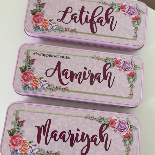 Three personalized pink metal nameplates or nameplates-style cases arranged vertically on a white surface. Each piece has floral rose illustrations in the corners, a decorative border, and a cursive name printed in the center: Latifah, Amiraah, and Mariyah.