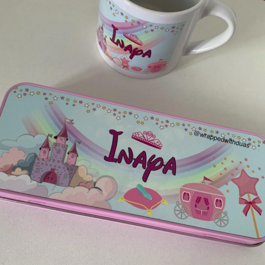 A rectangular metal pencil case with a pink border and colorful fantasy illustration, placed on a light surface next to a white ceramic mug. The pencil case features the word 'Inapa' in large purple letters with a tiara above it, along with a castle, carriage, and rainbow motifs.