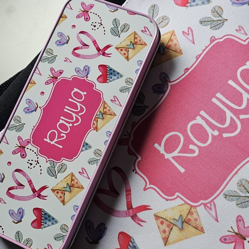A rectangular metal tin with a pink lid, decorated with a white background and colorful heart, ribbon, and envelope motifs, featuring the name 'Raya' in white script inside a pink label. It is placed diagonally on top of a matching printed card or box with the name 'Raya' in white script on a pink background.