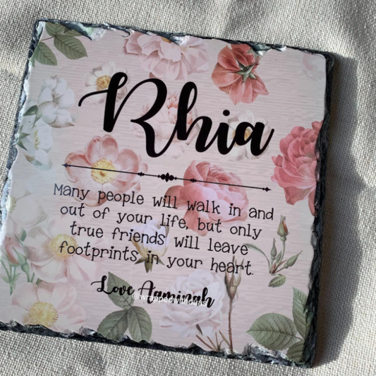 Personalized square slate plaque with floral border and the name 'Rhia' in large script, plus a quote and the name 'Lori Aaminah', photographed on a light fabric background.