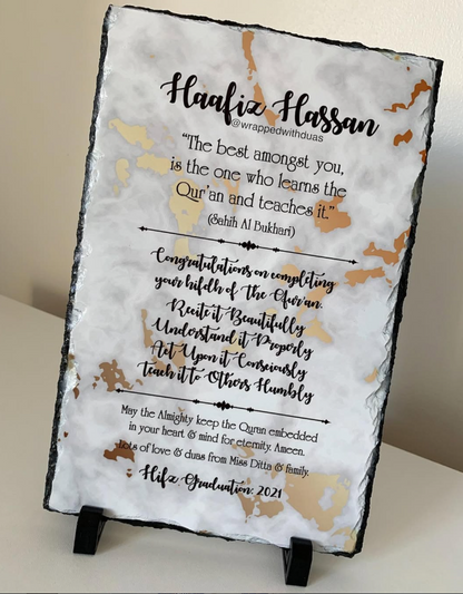 A square slate sign on a small black display stand, featuring a white marble-style background with gold abstract marbling and black text celebrating a Hafiz graduation with a Quran quote.
