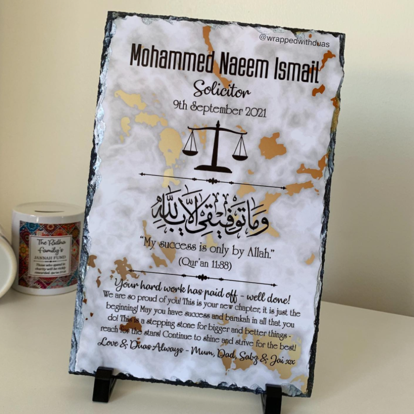 A personalized slate sign with a distressed black and white finish, featuring a gold foil background pattern, a black balance scale graphic, Arabic calligraphy, and a congratulatory message for a solicitor. The sign is displayed on a small black stand on a beige surface next to a jar candle.