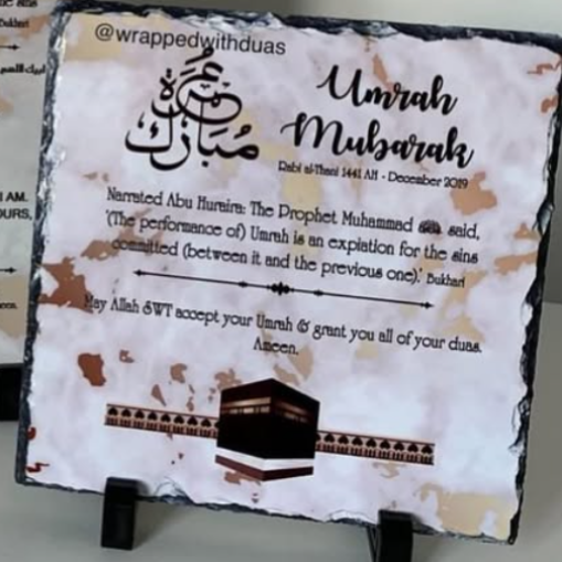 A square slate plaque with a black border, featuring Islamic calligraphy and text in Arabic and English, including 'Umrah Mubarak,' a hadith, and an illustration of the Kaaba. The plaque is displayed on a small black stand.