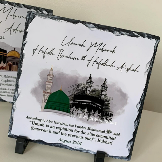 A square slate plaque on a small black stand, printed with a Ramadan farewell message, mosque illustrations, and a quote. A second plaque is partially visible behind it on a light beige surface.