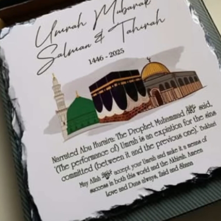 A printed card or plaque with Islamic commemorative text reading 'Umrah Mubarak Salman & Tahira 1446 - 2025' and an illustration of the Kaaba and mosque domes, photographed inside a box.