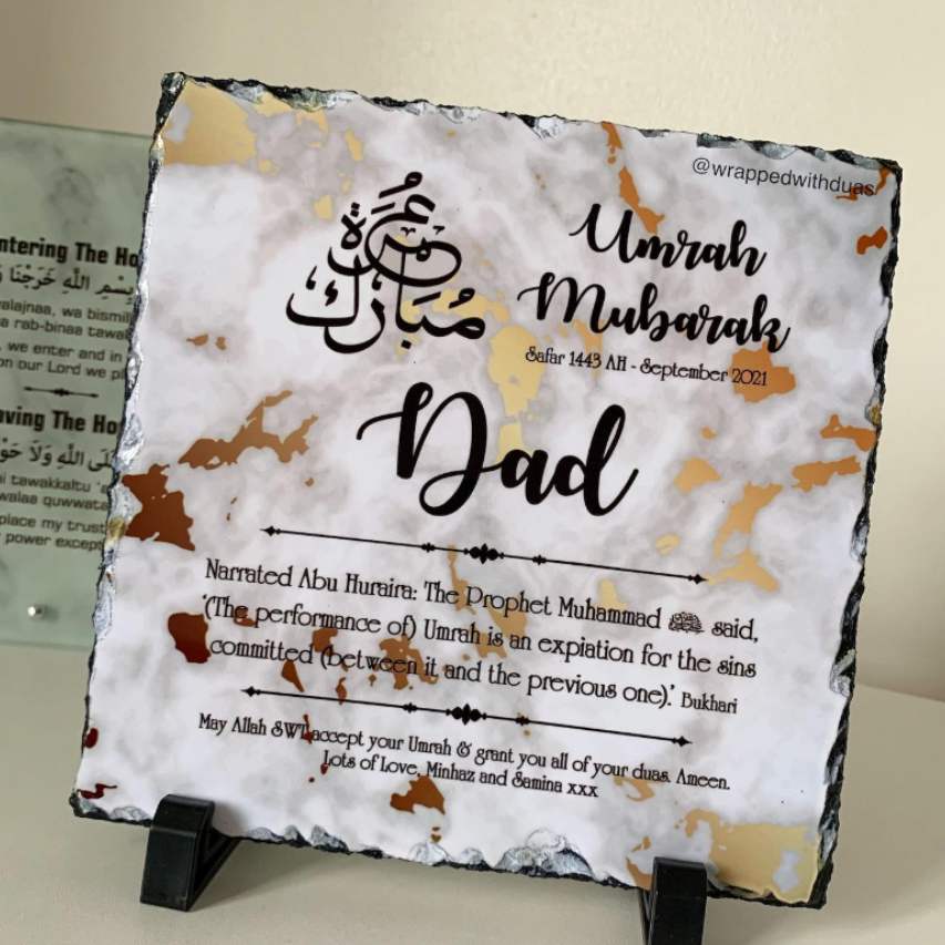 A rectangular slate plaque on a small black display stand, featuring engraved or printed Islamic text including 'Dad', 'Umrah Mubarak', dates, and prayer copy. The slate has a white base with brown and beige marbled veining and dark gray edges.