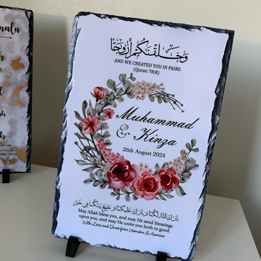 A personalized black slate sign with a white printed insert, featuring Arabic calligraphy, a floral wreath illustration with pink and red roses, and the names 'Muhammad & Kenza' with a date. The sign is displayed on a tabletop with a small black stand.