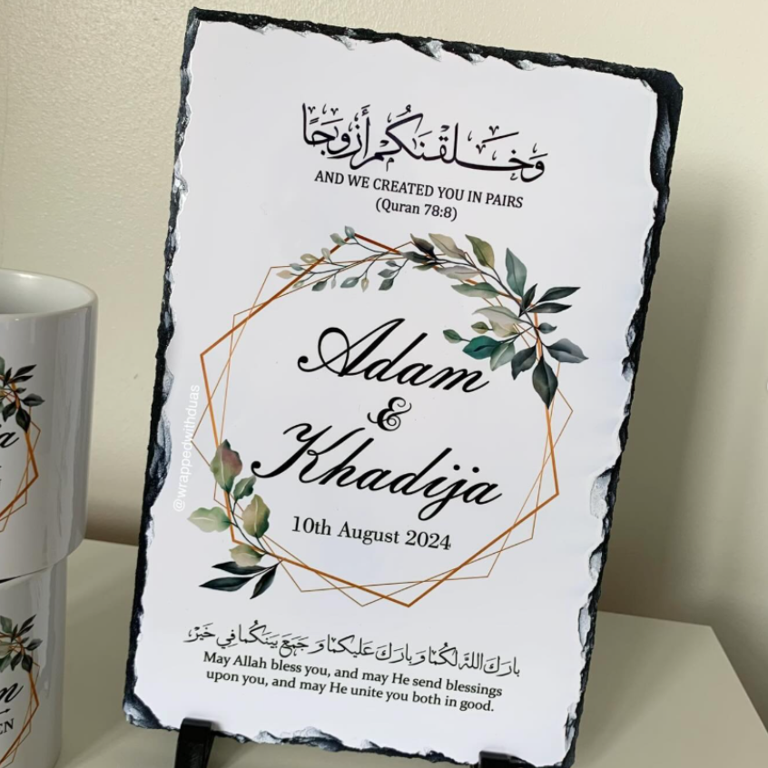 Personalized black slate wedding sign with a white printed insert, featuring Arabic and English text, a geometric gold line design, and green leaf illustrations. The sign is displayed on a tabletop with a matching ceramic mug partially visible on the left.