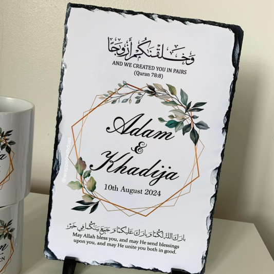 Personalized black slate wedding sign with a white printed insert, featuring Arabic and English text, a geometric gold line design, and green leaf illustrations. The sign is displayed on a tabletop with a matching ceramic mug partially visible on the left.