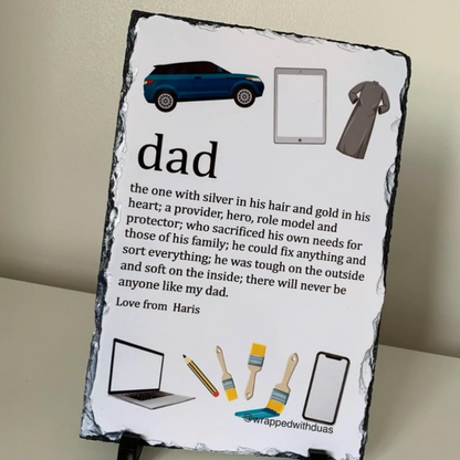 A personalized slate sign featuring a 'dad' dictionary definition with illustrations of things a dad might do, displayed on a small black stand against a beige wall.