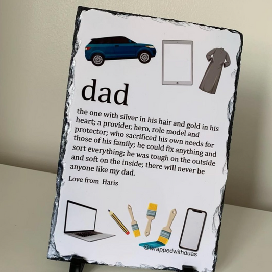 A personalized slate sign featuring a 'dad' dictionary definition with illustrations of things a dad might do, displayed on a small black stand against a beige wall.