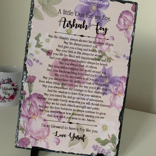 A square decorative plaque or sign with a black slate border, featuring a floral design in purple and green, with a printed poem titled 'A Little Dua for my Foy' and the name 'Aishah Foy' in large script. The sign is displayed on a small black stand next to a white ceramic mug with a matching design.