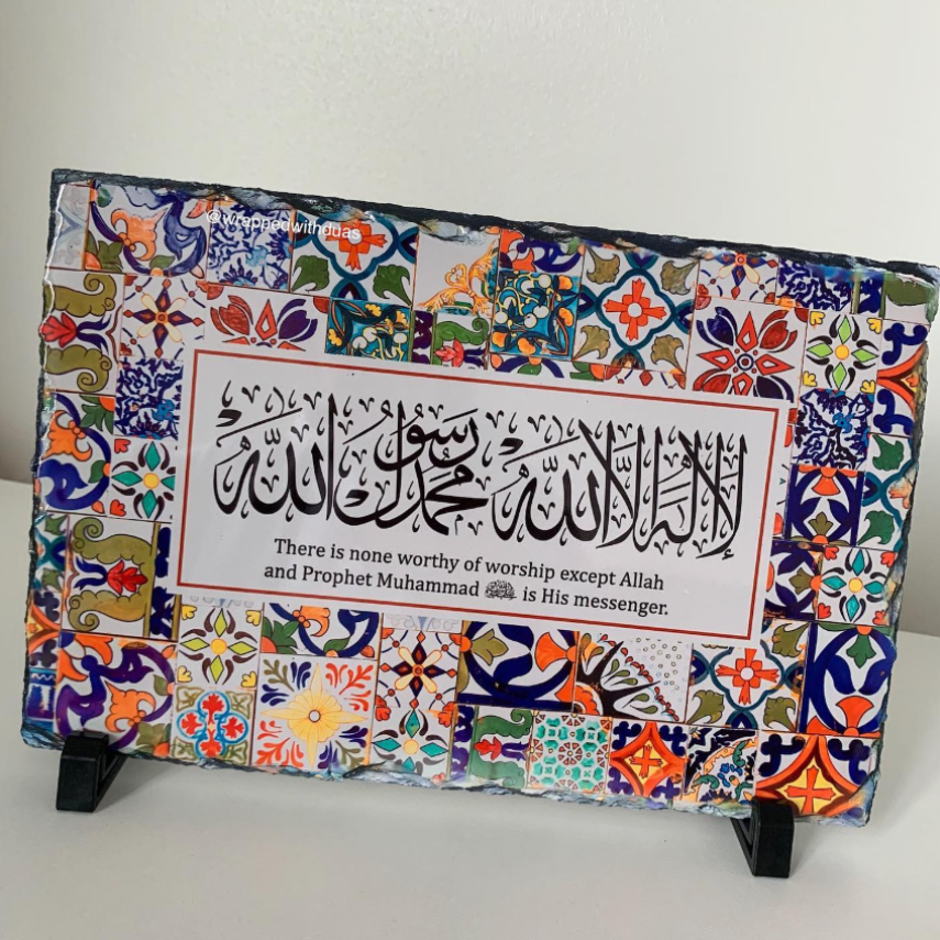 A rectangular decorative plaque with colorful Islamic calligraphy and a quote, mounted on a black display stand against a plain light wall.