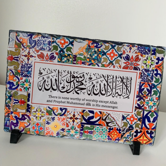 A rectangular decorative plaque with colorful Islamic calligraphy and a quote, mounted on a black display stand against a plain light wall.