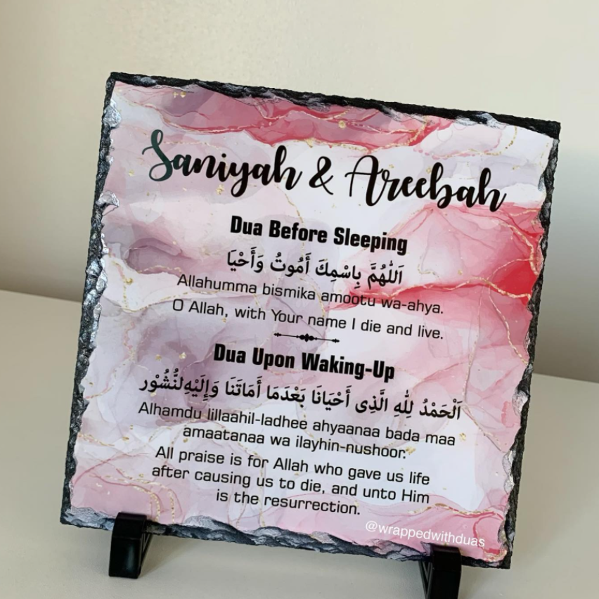 A square slate plaque on a small black stand, featuring a pink and white marbled background with black text reading 'Saniyah & Areebah' and dua prayer copy for before sleeping and upon waking. The plaque is photographed indoors against a light neutral wall.