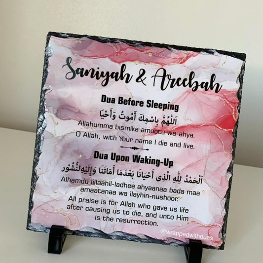 A square slate plaque on a small black stand, featuring a pink and white marbled background with black text reading 'Saniyah & Areebah' and dua prayer copy for before sleeping and upon waking. The plaque is photographed indoors against a light neutral wall.